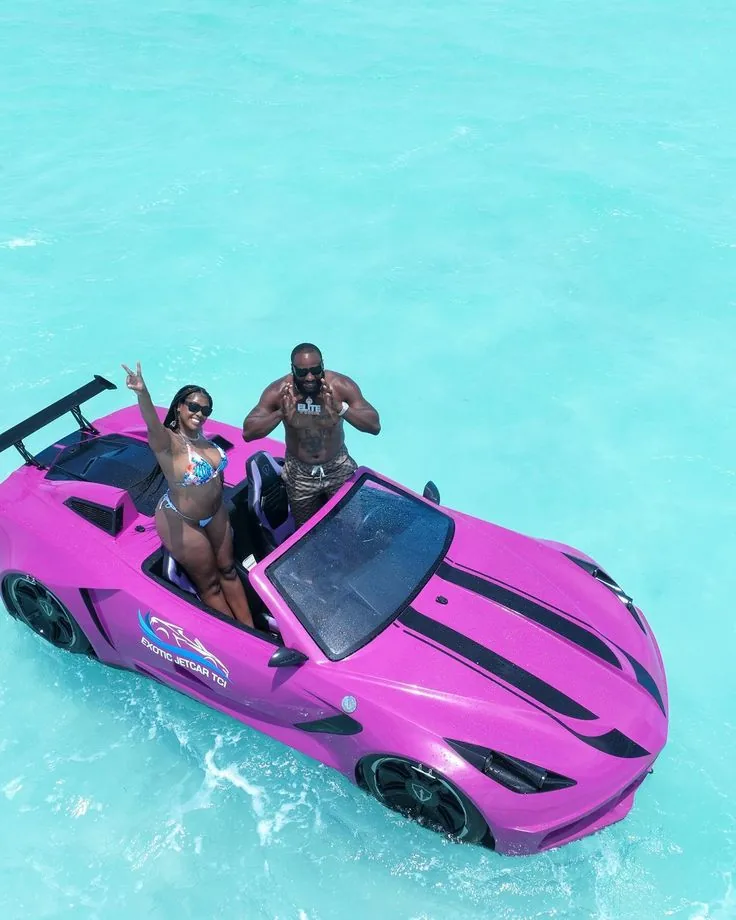 Two people sitting side by side in a small jet-powered water car, riding over calm turquoise ocean water near the Zanzibar coastline. The sea is clear and blue, with gentle waves and a sunny tropical sky in the background.